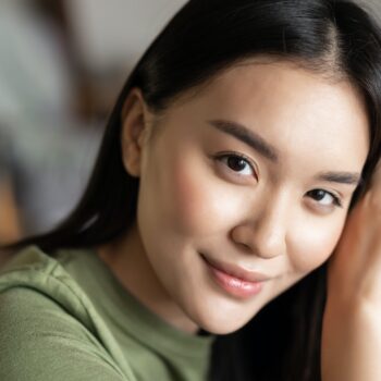 Close up portrait of tender asian girl with glowing natural skin, smiling sensual at camera, sitting