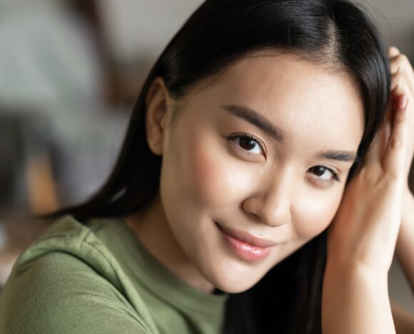 Close up portrait of tender asian girl with glowing natural skin, smiling sensual at camera, sitting