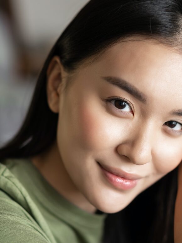 Close up portrait of tender asian girl with glowing natural skin, smiling sensual at camera, sitting