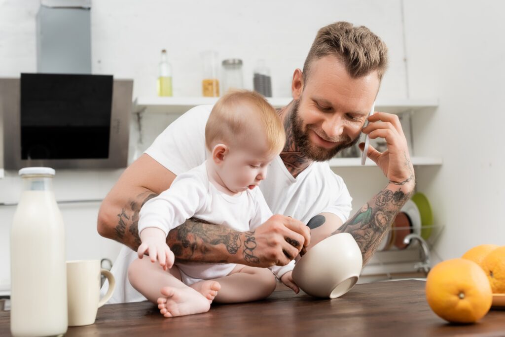 young tattooed man talking on smartphone near infant son sitting on kitchen table during breakfast